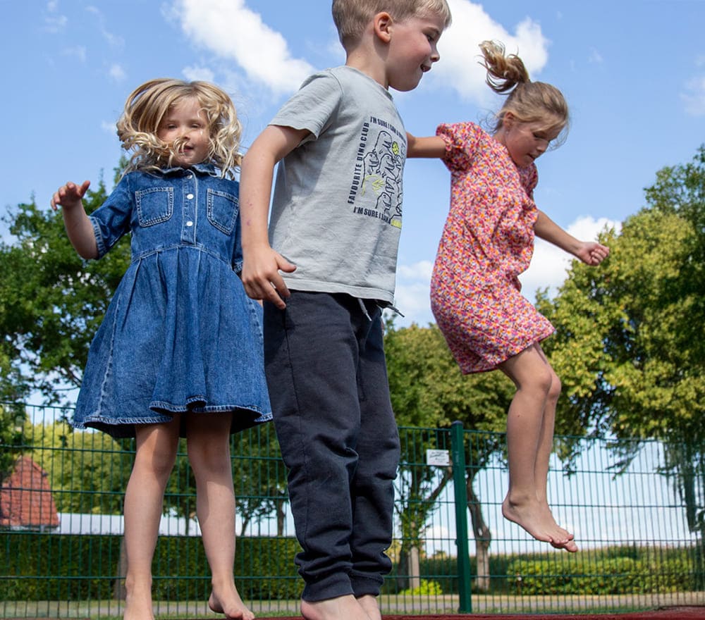 Drei kleine Kinder springen an einem sonnigen Tag barfuß draußen herum und genießen die Freiheit, die man bei der Reittherapie für Kinder oft erlebt. Zwei Mädchen tragen Kleider, ein Junge trägt ein T-Shirt und eine Hose. Im Hintergrund sind grüne Bäume und ein Metallzaun zu sehen.