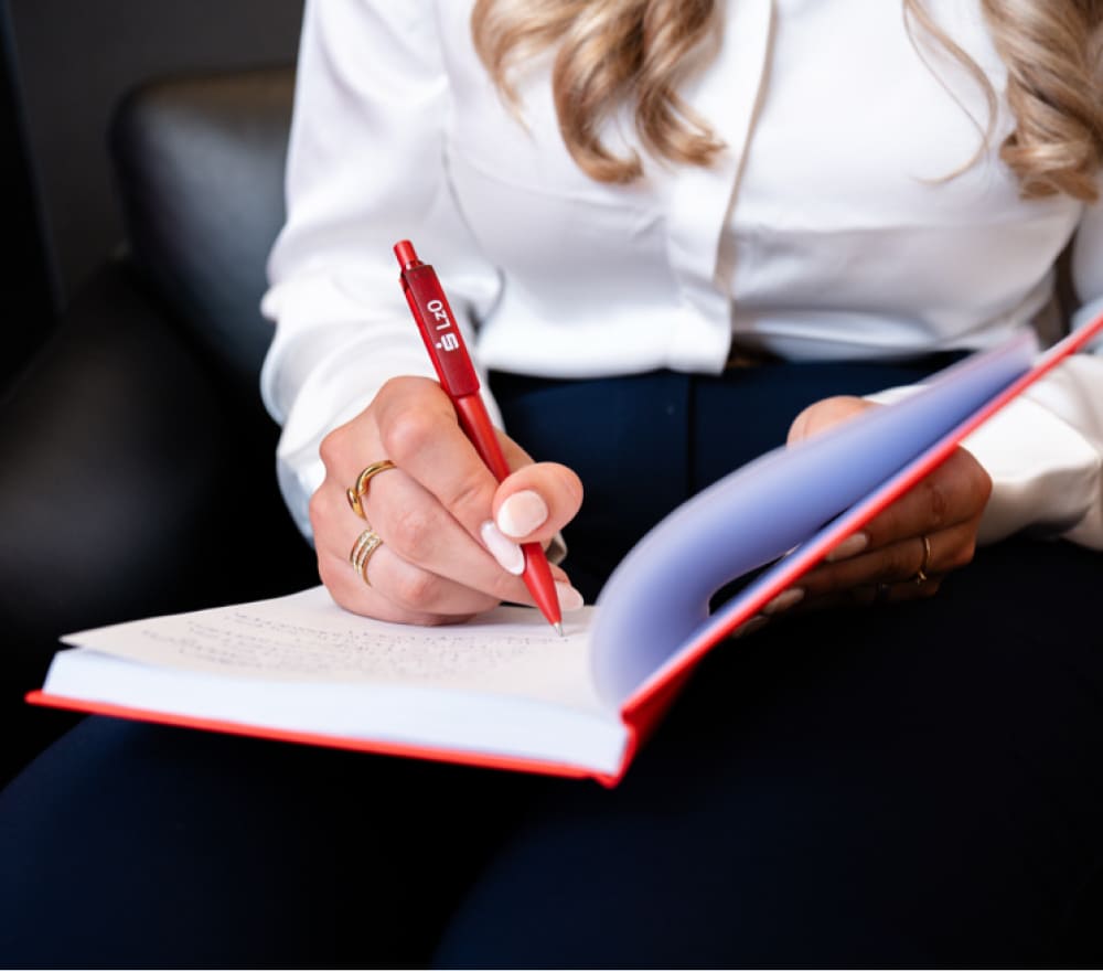 Eine Person mit langen, welligen Haaren, die ein weißes Hemd trägt, sitzt auf einem dunklen Stuhl und schreibt mit einem roten Stift in ein Notizbuch, während sie sorgfältig ihre Finanzen zum Ausbildungsstart plant.