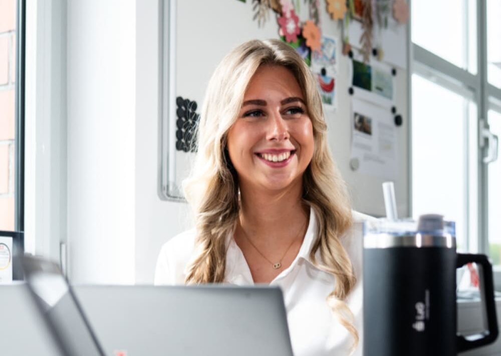 Eine lächelnde Frau mit langen blonden Haaren sitzt an einem Schreibtisch mit einem Dell-Laptop und einem großen schwarzen Becher und macht sich bereit, ihre Finanzen in den Griff zu bekommen, während sie sich auf den Ausbildungsstart vorbereitet. Hinter ihr sind auf einem Whiteboard verschiedene Notizen, Fotos und Dekorationen zu sehen. Sonnenlicht strömt durch die Fenster in der Nähe herein.