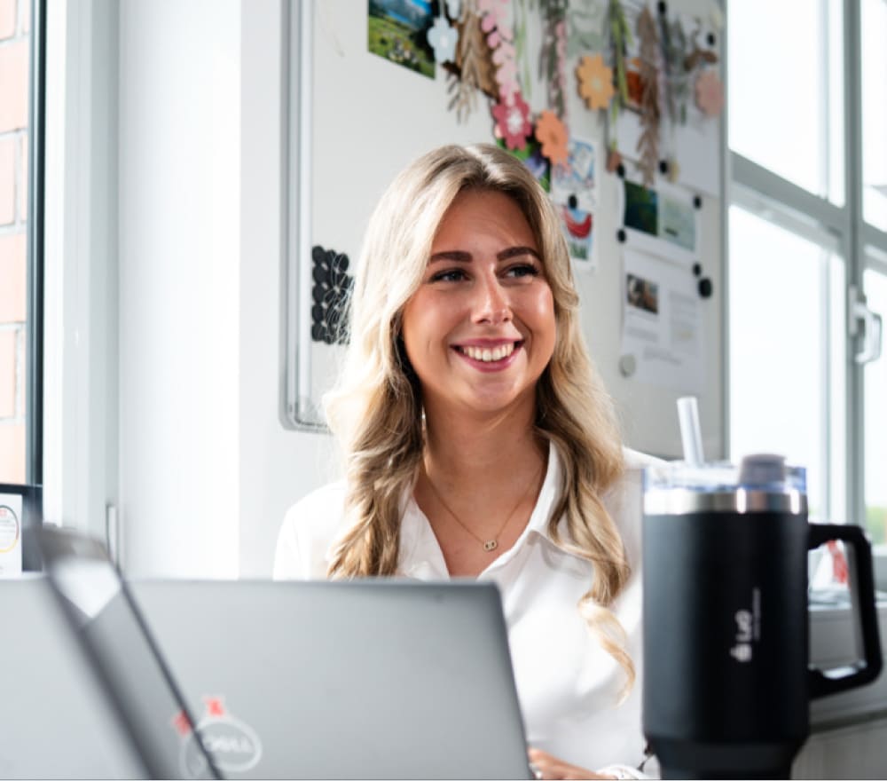 Eine lächelnde Frau mit langen blonden Haaren sitzt an einem Schreibtisch mit einem Dell-Laptop und einem großen schwarzen Becher und macht sich bereit, ihre Finanzen in den Griff zu bekommen, während sie sich auf den Ausbildungsstart vorbereitet. Hinter ihr sind auf einem Whiteboard verschiedene Notizen, Fotos und Dekorationen zu sehen. Sonnenlicht strömt durch die Fenster in der Nähe herein.