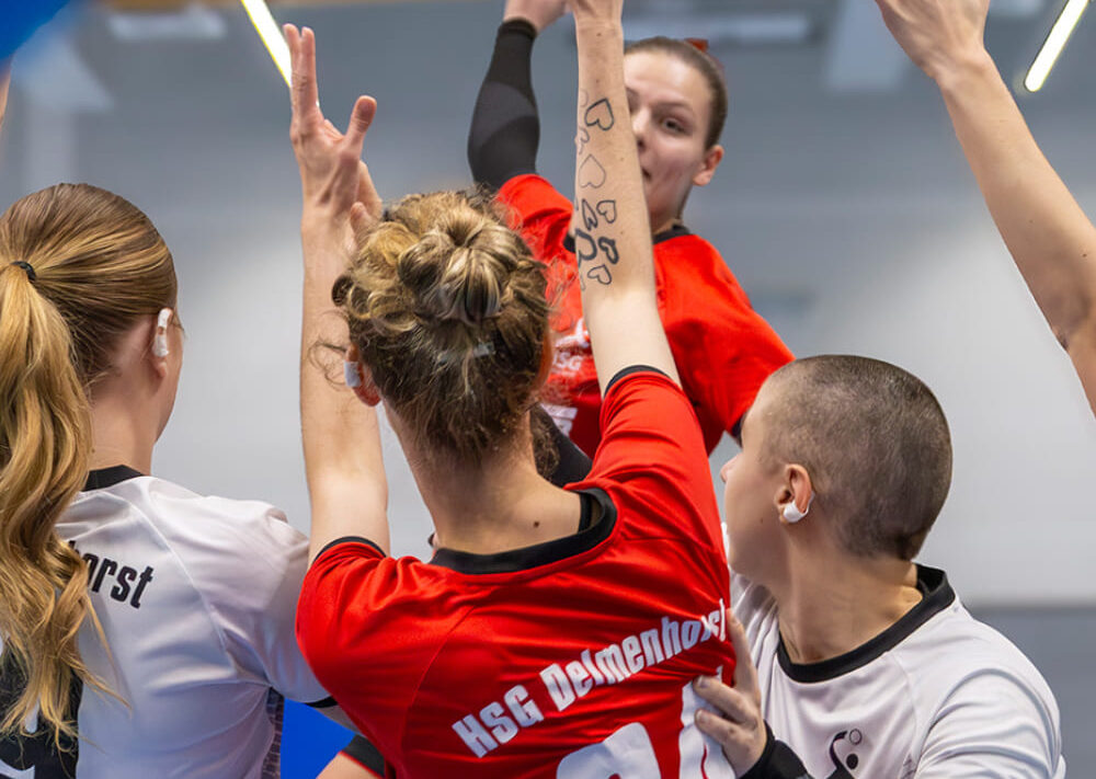 Eine Handballerin in einem roten Trikot springt hoch, um den Ball zu werfen, dicht umringt von drei gegnerischen Spielerinnen in weißen Trikots, die alle versuchen, ihren Wurf während eines intensiven Spiels zu blocken – ein Beispiel für die Art von Teamwork und Koordination, die oft durch Aktivitäten wie die Reittherapie für Kinder gefördert wird.