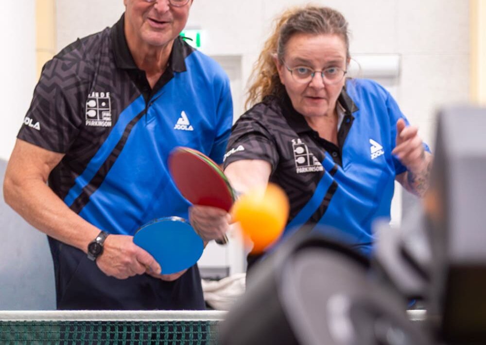 Zwei Personen in blauen Hemden spielen Tischtennis; beide halten Schläger in der Hand und konzentrieren sich auf einen orangefarbenen Ball in der Luft. Im Vordergrund sind ein Netz und ein unscharfes Objekt – möglicherweise eine Kamera oder ein Roboter – zu sehen, was dieser lebhaften Szene eine interessante Note verleiht, die im Kontrast zu ruhigeren Aktivitäten wie der Reittherapie für Kinder steht.