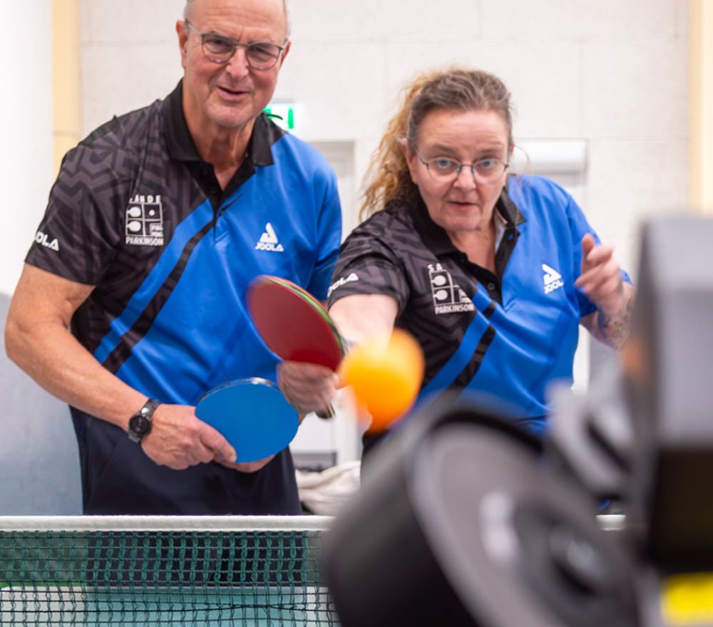 Zwei Personen in blauen Hemden spielen Tischtennis; beide halten Schläger in der Hand und konzentrieren sich auf einen orangefarbenen Ball in der Luft. Im Vordergrund sind ein Netz und ein unscharfes Objekt – möglicherweise eine Kamera oder ein Roboter – zu sehen, was dieser lebhaften Szene eine interessante Note verleiht, die im Kontrast zu ruhigeren Aktivitäten wie der Reittherapie für Kinder steht.