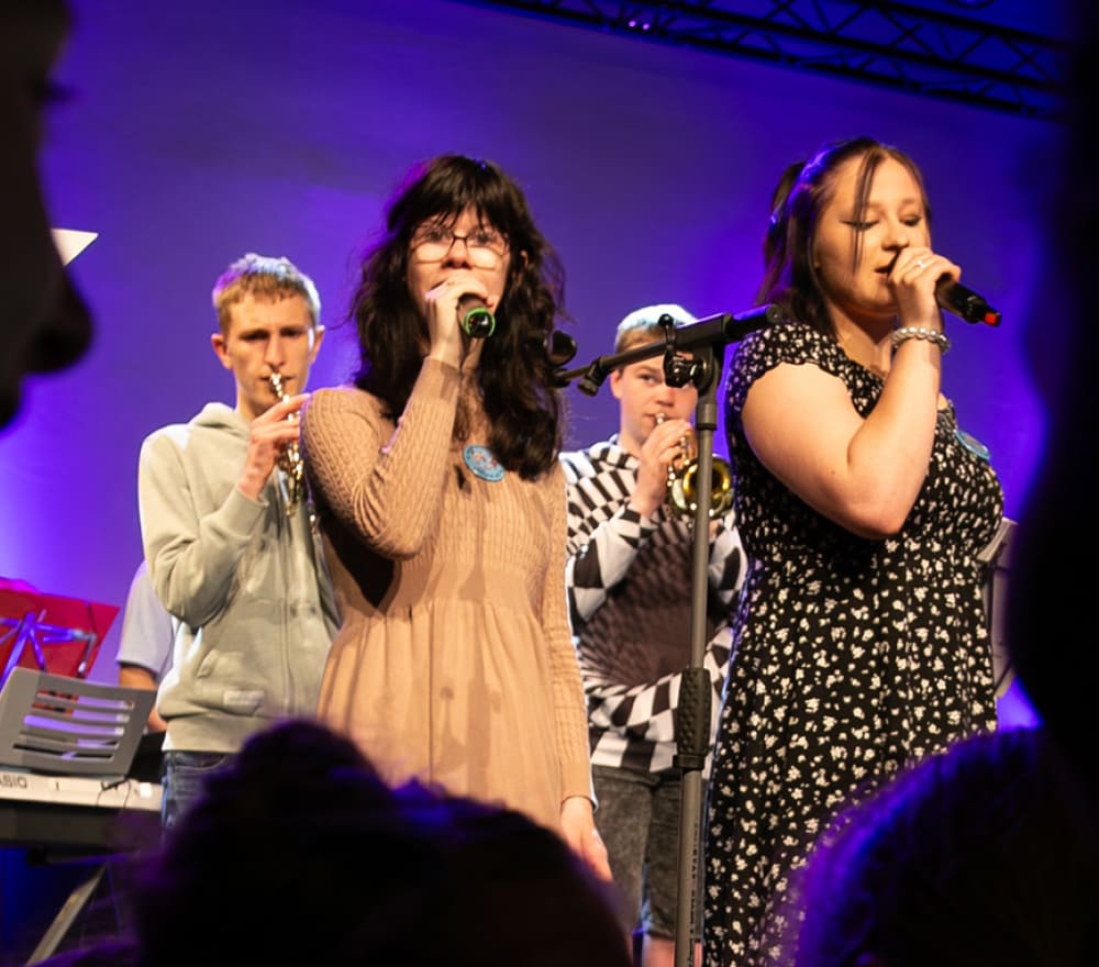 Vier junge Menschen treten auf der Bühne auf; zwei singen vorne in Mikrofone, während zwei im Hintergrund Trompete spielen. Die Bühnenbeleuchtung taucht den Raum in violette Farbtöne, und im Vordergrund sind die Silhouetten der Zuschauer zu erkennen. Die Veranstaltung soll das Bewusstsein für „Reittherapie für Kinder“ schärfen und verbindet Musik mit einem guten Zweck.