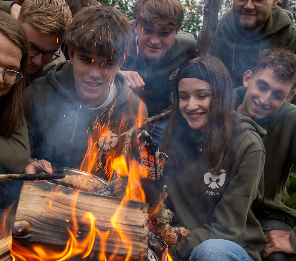 Eine Gruppe von Jugendlichen in grünen Kapuzenpullis sitzt eng beieinander, lächelt und beobachtet ein Lagerfeuer mit hellen Flammen und fliegenden Funken. Auf einem der Kapuzenpullis steht der Name „Anna“. Die Szene, die an gemeinsame Momente nach den Sitzungen der Reittherapie für Kinder erinnert, spielt sich vor dem Hintergrund von Bäumen ab, die im warmen Abendlicht baden.