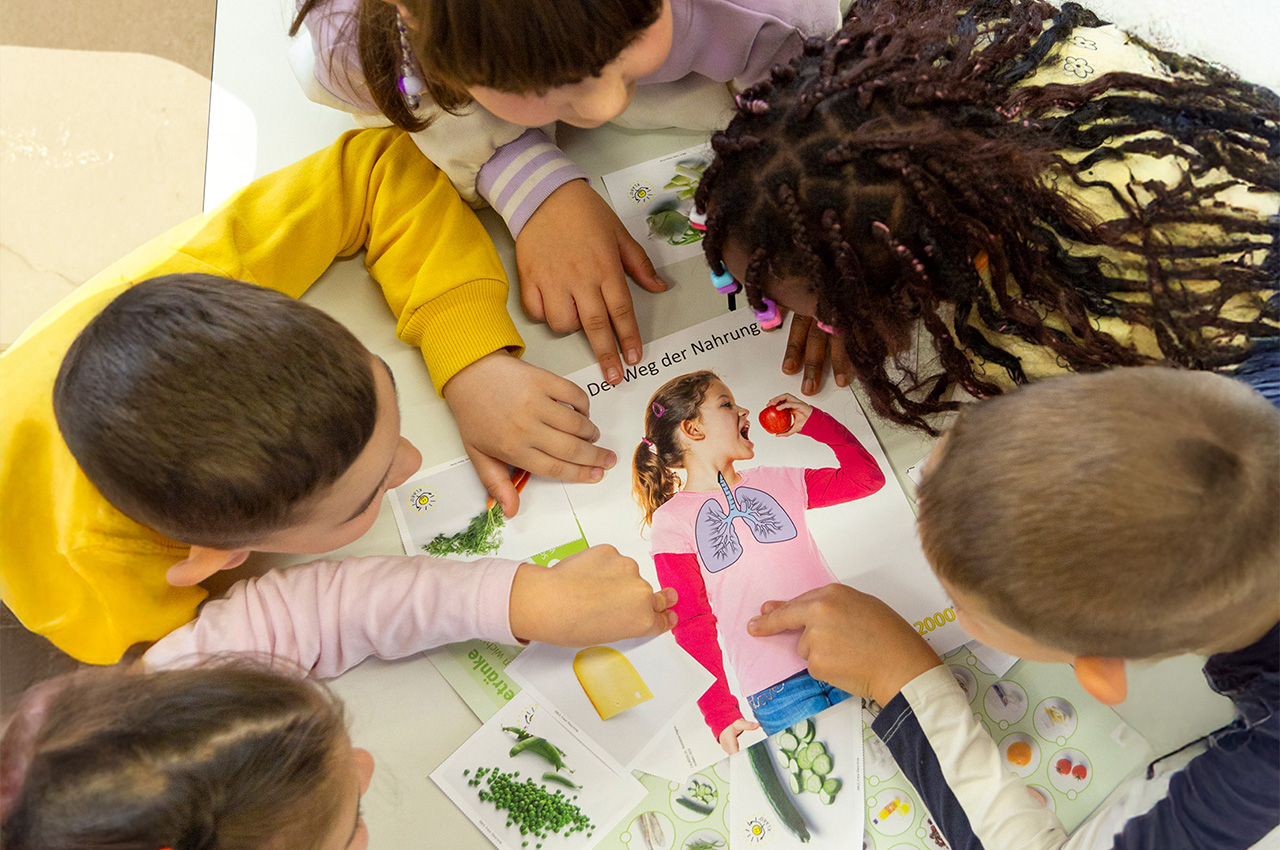 Fünf Kinder sitzen um einen Tisch herum und zeigen auf ein buntes Lernplakat mit Abbildungen von Lebensmitteln, menschlichen Organen und deutschen Texten. Die Kinder sind begeistert dabei und beschäftigen sich gemeinsam mit dem Plakat, wobei sie im Unterricht Themen wie die Digitalisierung erkunden – vielleicht stellen sie dabei sogar einen Bezug zu lokalen Organisationen wie dem Feuerwehr-Spielmannszug Cloppenburg her.