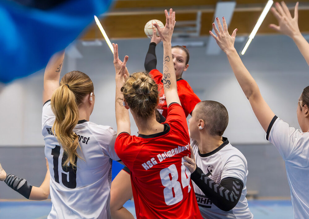 Eine Handballerin in einem roten Trikot des VSK Bungerhof Handball springt hoch, um den Ball zu werfen, während sie während eines intensiven Hallenspiels von vier gegnerischen Spielerinnen in weißen Trikots umringt und geblockt wird.