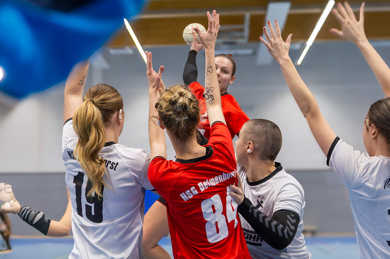 Eine Handballerin in einem roten Trikot des VSK Bungerhof Handball springt hoch, um den Ball zu werfen, während sie während eines intensiven Hallenspiels von vier gegnerischen Spielerinnen in weißen Trikots umringt und geblockt wird.