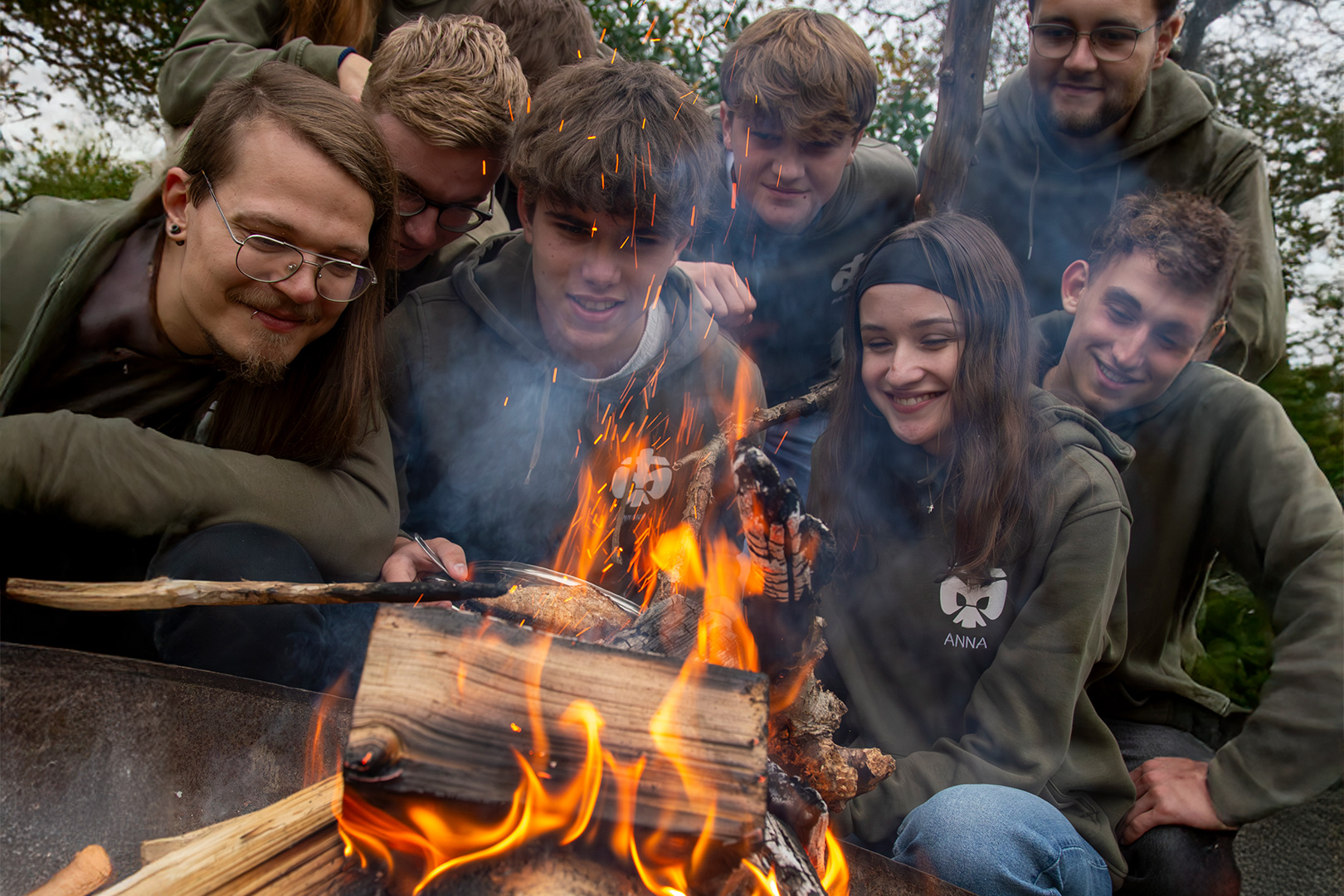 Eine Gruppe junger Leute in Kapuzenpullis sitzt um ein Lagerfeuer herum, brät Essen am Spieß, lächelt und genießt die Natur – ganz ähnlich wie die Kameradschaft bei einem Treffen des Männerchors „HatMannTöne Großenkneten“, wo Gesang und Gemeinschaftsgeist durch Vereinsförderung gepflegt werden.