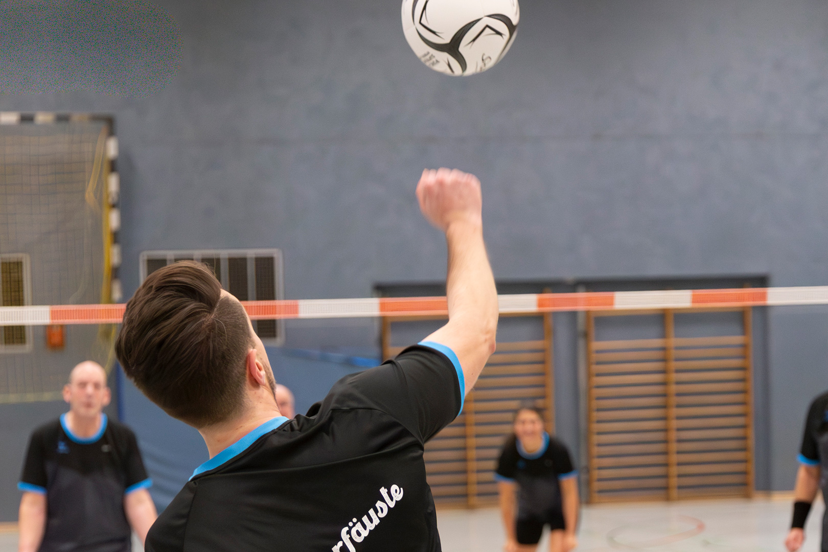Eine Person in einem schwarzen Sporttrikot schlägt einen Volleyball in einer Halle auf, im Hintergrund sind Teamkollegen und ein Netz zu sehen – ein Bild, das den Geist der Inklusion einfängt, der oft bei Veranstaltungen wie dem Theaterfestival Inklusion MUTIK Förderungen gefeiert wird.