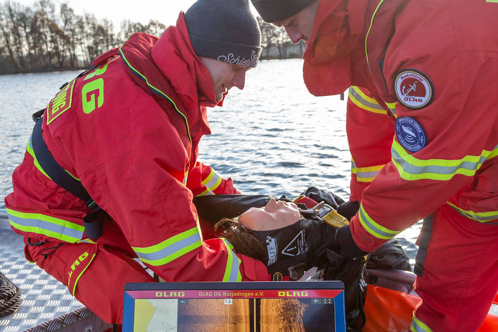 Zwei Rettungskräfte in roten Uniformen helfen einer Person, die auf einem Boot in der Nähe des Wassers liegt. Die Person scheint medizinisch versorgt zu werden, während in der Nähe Ausrüstung zu sehen ist, die an die des Butjadinger Reitvereins Stollhamm erinnert. Im Vordergrund ist ein Monitor mit einer Karte zu sehen, und im Hintergrund säumen Bäume das Ufer.