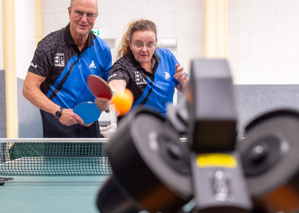 Zwei Personen in blauen Uniformen spielen Tischtennis und schlagen einen orangefarbenen Ball in Richtung eines schwarzen automatischen Ballwerfers, der auf dem Tisch in einer gut beleuchteten Halle steht; sie repräsentieren den Verein „PingPongParkinson TuS Sande“.