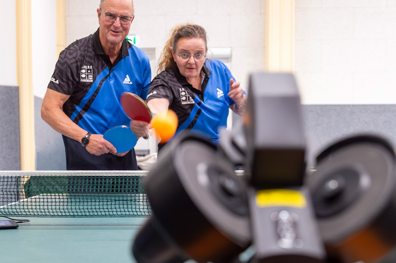 Zwei Personen in blauen Uniformen spielen Tischtennis und schlagen einen orangefarbenen Ball in Richtung eines schwarzen automatischen Ballwerfers, der auf dem Tisch in einer gut beleuchteten Halle steht; sie repräsentieren den Verein „PingPongParkinson TuS Sande“.