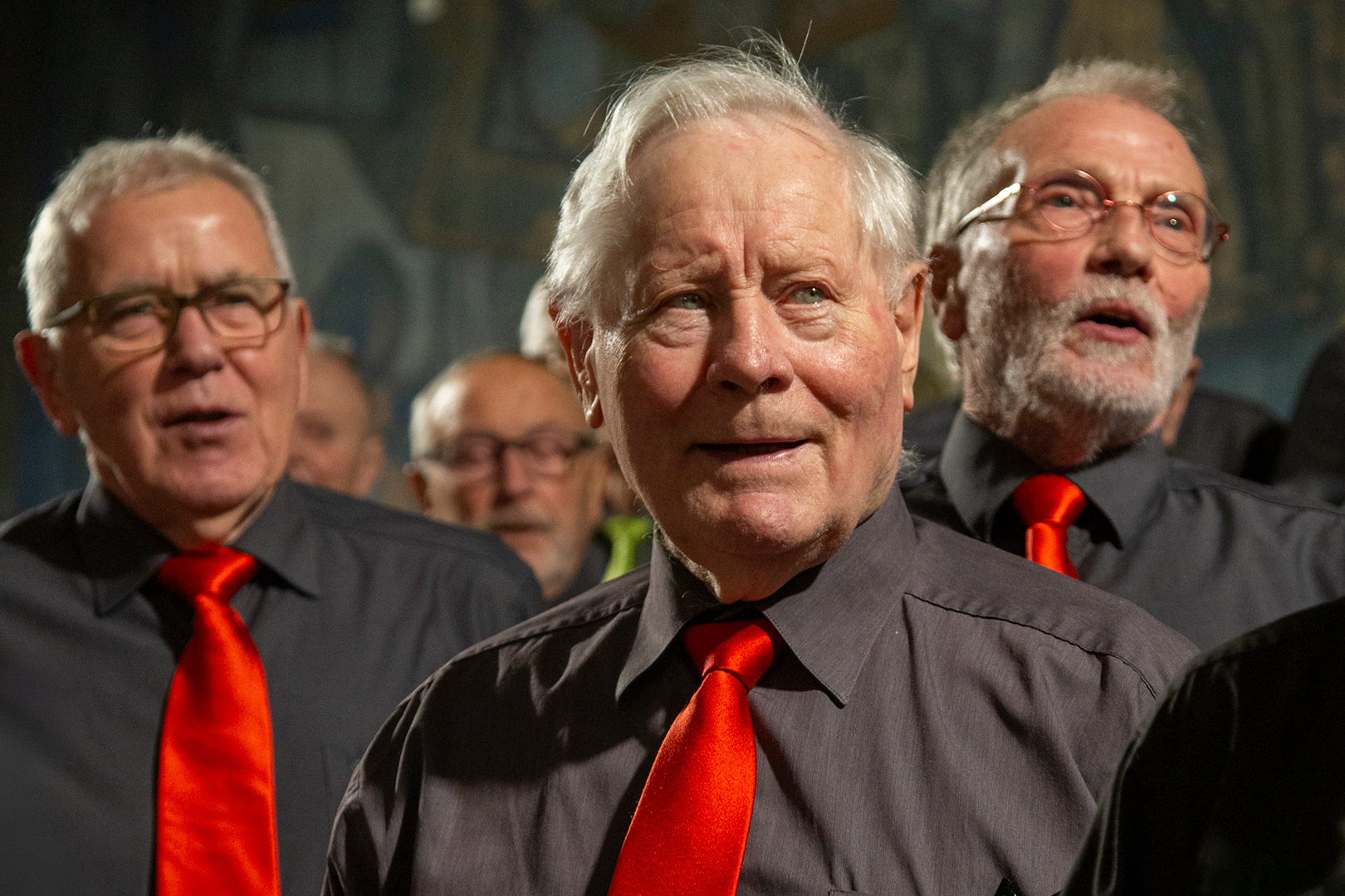 Eine Nahaufnahme einer Gruppe älterer Männer, die beim Theaterfestival „Inklusion“ im Chor singen. Sie tragen dunkelgraue Hemden mit leuchtend roten Krawatten. Im Mittelpunkt steht ein Mann mit weißem Haar in der Mitte, flankiert von anderen Männern mit Brille, die alle den Mund leicht geöffnet haben.