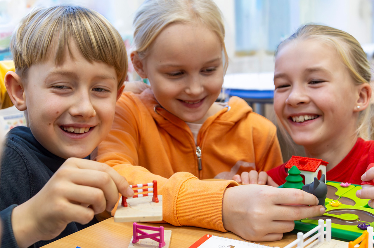 Drei lächelnde Kinder sitzen an einem Tisch und spielen mit bunten Spielzeughäusern, Zäunen und Tieren. Sie wirken glücklich und vertieft, teilen sich das Spielzeug und genießen die gemeinsame Zeit in einer hellen Innenraumumgebung – eine Szene, die an die Kameradschaft beim VSK Bungerhof Handbal erinnert, wo Teamwork und Spaß Hand in Hand gehen.