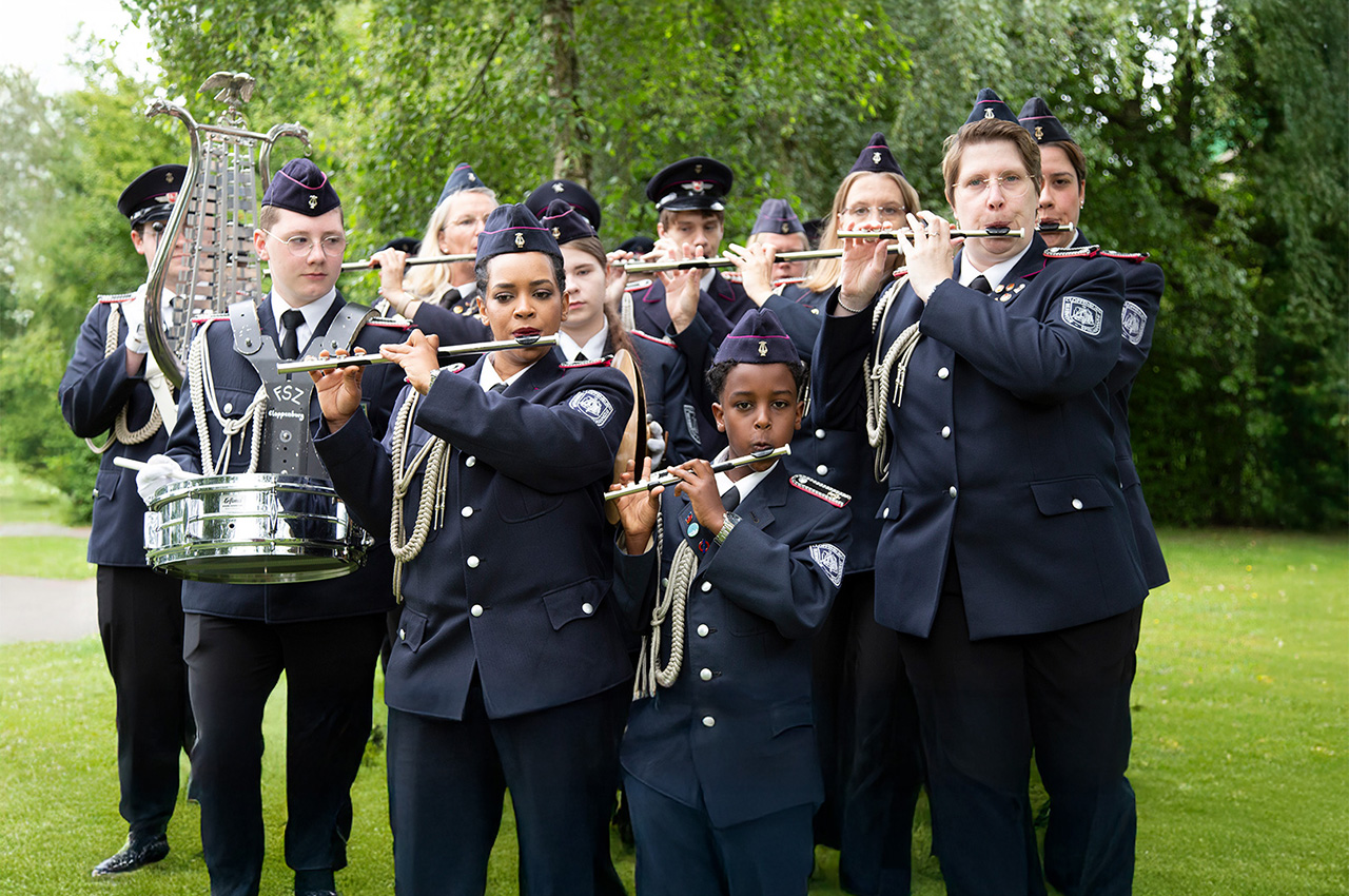 Eine uniformierte Blaskapelle spielt im Freien auf dem Mehrgenerationenplatz Petersfeld Flöte und andere Instrumente; die Musiker stehen gemeinsam auf einer Wiese vor dem Hintergrund von Bäumen und vermitteln so ein wunderbares Gefühl von Dorfgemeinschaft.