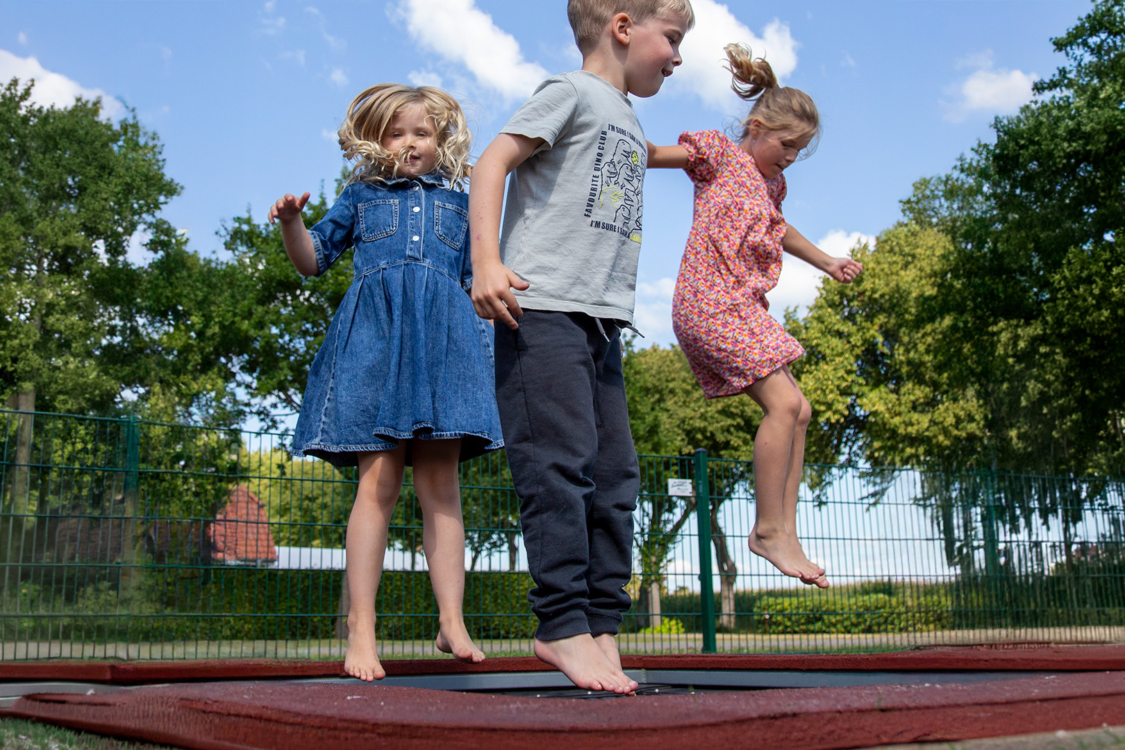 Drei kleine Kinder spielen und hüpfen barfuß auf einem Trampolin im Freien, umgeben von grünen Bäumen und einem Metallzaun, unter einem strahlend blauen Himmel mit einigen Wolken – eine lebhafte Szene, die an die fröhliche Stimmung im Ferienlager der DPSG Füchtel Pfadfinder Jugendarbeit erinnert.