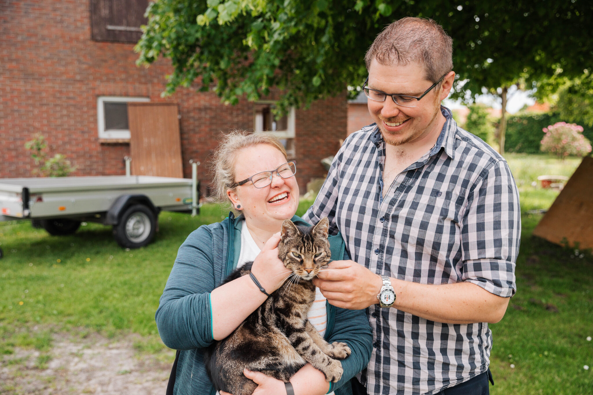 Zwei lächelnde Erwachsene stehen draußen und halten eine getigerte Katze im Arm; im Hintergrund sind ein Wohnwagen und ein Backsteingebäude zu sehen. Die Szene ist sonnig und grün, und beide Menschen sehen glücklich aus, während sie die Katze streicheln.