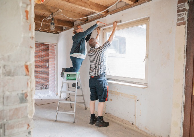 Zwei Personen arbeiten gemeinsam an einer Hausrenovierung: Die eine steht auf einer Trittleiter, die andere auf dem Boden, und beide messen oder justieren etwas über einem Fenster in einem noch nicht ganz fertiggestellten Raum.
