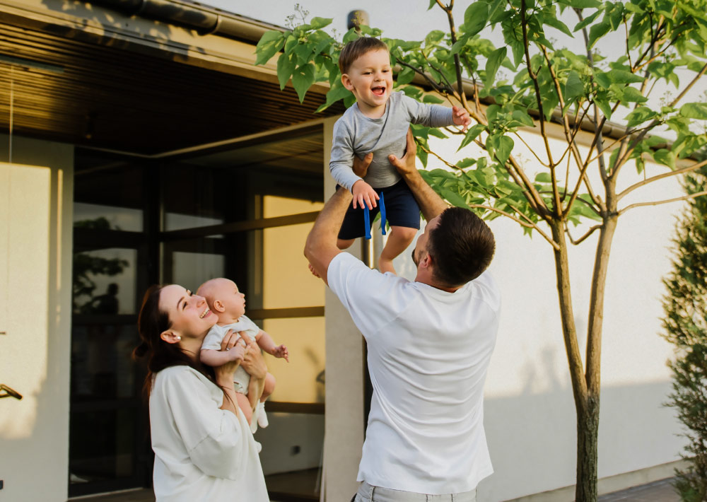 Ein Mann hebt einen lächelnden kleinen Jungen in die Luft, während eine Frau ein Baby im Arm hält. Sie befinden sich draußen in der Nähe eines Hauses, stehen unter einem kleinen Baum und genießen gemeinsam einen sonnigen Tag.