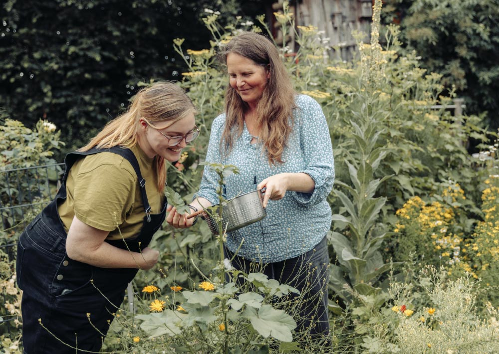 Zwei Frauen, die lächelnd gemeinsam im Freien gärtnern. Die eine hält ein Metall-Sieb in der Hand, während die andere zwischen hohem grünem Laub und gelben Blumen die Pflanzen begutachtet.