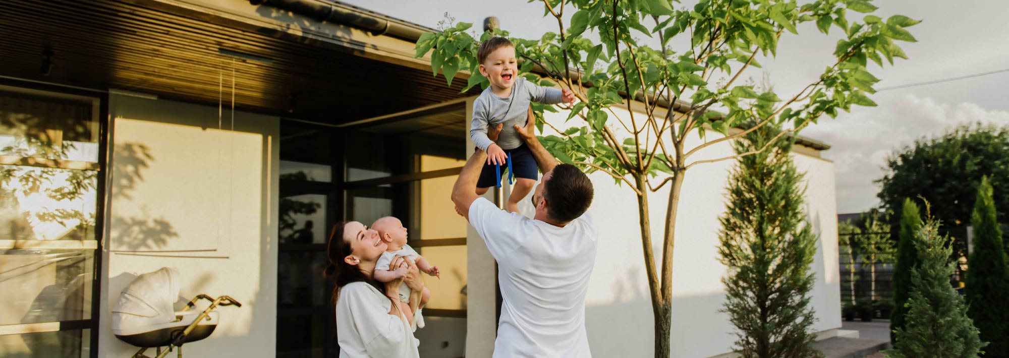 Ein Mann hebt einen lächelnden kleinen Jungen in die Luft, während eine Frau in der Nähe ein Baby im Arm hält. Sie befinden sich vor einem modernen Haus, umgeben von Grünflächen und Bäumen, und genießen gemeinsam als Familie einen sonnigen Tag.