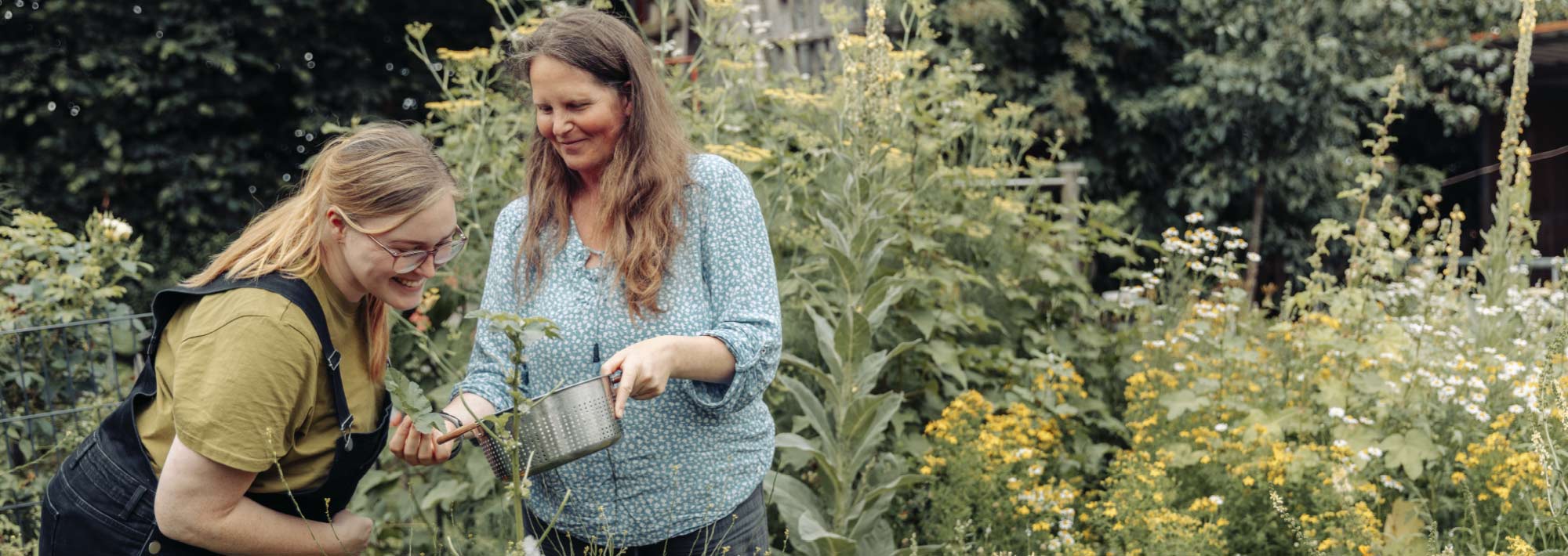 Zwei Frauen stehen in einem üppigen Garten, lächeln und pflücken gemeinsam Kräuter oder Pflanzen. Die eine hält eine Metallschüssel in der Hand, während die andere nach den Pflanzen greift. Sie sind von Grünpflanzen und Blumen umgeben.