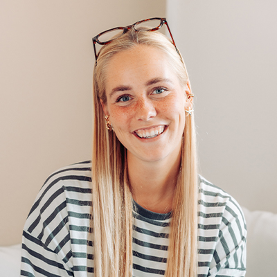 Eine lächelnde Frau mit langen blonden Haaren, die ein gestreiftes Shirt trägt und deren Brille auf dem Kopf sitzt, sitzt drinnen vor einer hellen Wand.