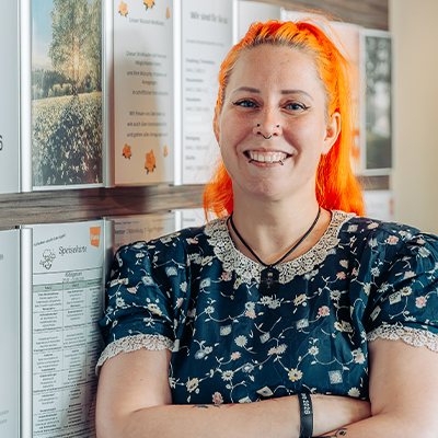 Eine Person mit leuchtend orangefarbenen Haaren und einem geblümten Kleid steht lächelnd mit verschränkten Armen vor einer Wand, die mit Papieren und Plakaten bedeckt ist.