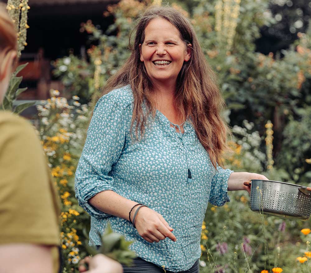 Eine Frau mit langen braunen Haaren, die ein blau gemustertes Hemd trägt, lächelt, während sie in einem üppigen, von gelben Blumen umgebenen Garten ein Metall-Sieb in der Hand hält. Im Vordergrund ist eine weitere Person teilweise zu sehen.