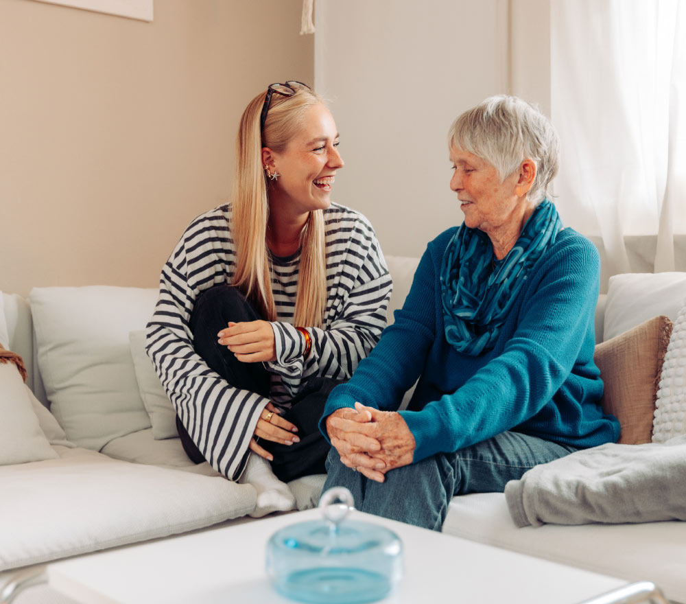 Eine junge Frau und eine ältere Frau sitzen auf einem Sofa, lächeln und unterhalten sich in einem hellen, gemütlichen Wohnzimmer.