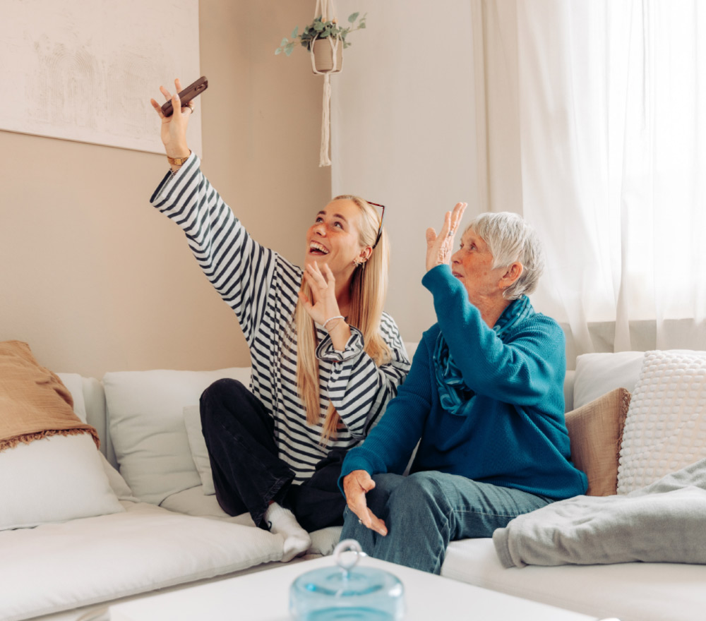 Zwei Frauen sitzen auf einem Sofa, lächeln und winken, während die jüngere Frau ihr Handy hochhält, um ein Selfie zu machen. Der Raum wirkt gemütlich mit sanfter Beleuchtung, Kissen und einer Decke.