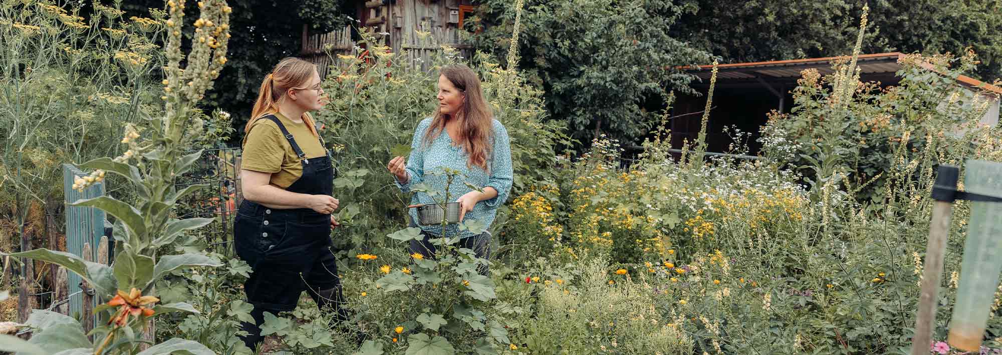 Zwei Frauen stehen in einem üppigen, grünen Garten voller Blumen und hoher Pflanzen und unterhalten sich. Eine von ihnen hält einen Korb in der Hand, und im Hintergrund sind ein Schuppen und Bäume zu sehen.