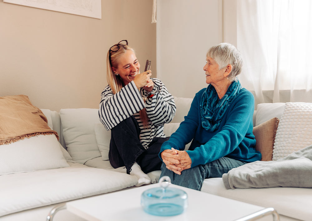 Eine junge Frau und eine ältere Frau sitzen auf einem weißen Sofa, lächeln und unterhalten sich. Die jüngere Frau hält ein Smartphone in der Hand, und beide wirken glücklich und entspannt in einem gemütlichen, hell erleuchteten Wohnzimmer.