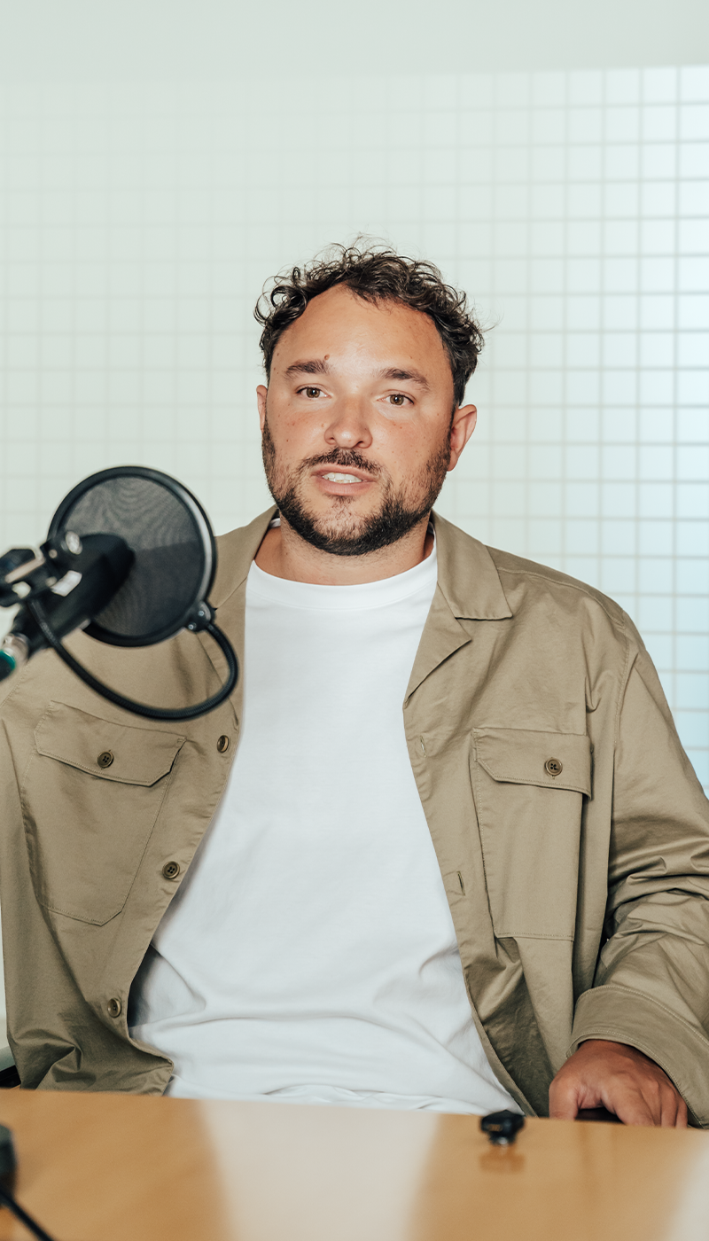 Ein Mann mit kurzen Locken sitzt an einem Tisch vor einem Mikrofon; er trägt eine beige Jacke über einem weißen Hemd und steht vor einem weißen Fliesenhintergrund.