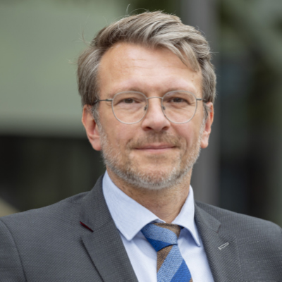 Ein Mann mittleren Alters mit grauen Haaren, Bart und Brille, der ein Sakko, ein hellblaues Hemd und eine gestreifte Krawatte trägt und im Freien vor einem unscharfen Gebäude im Hintergrund steht.