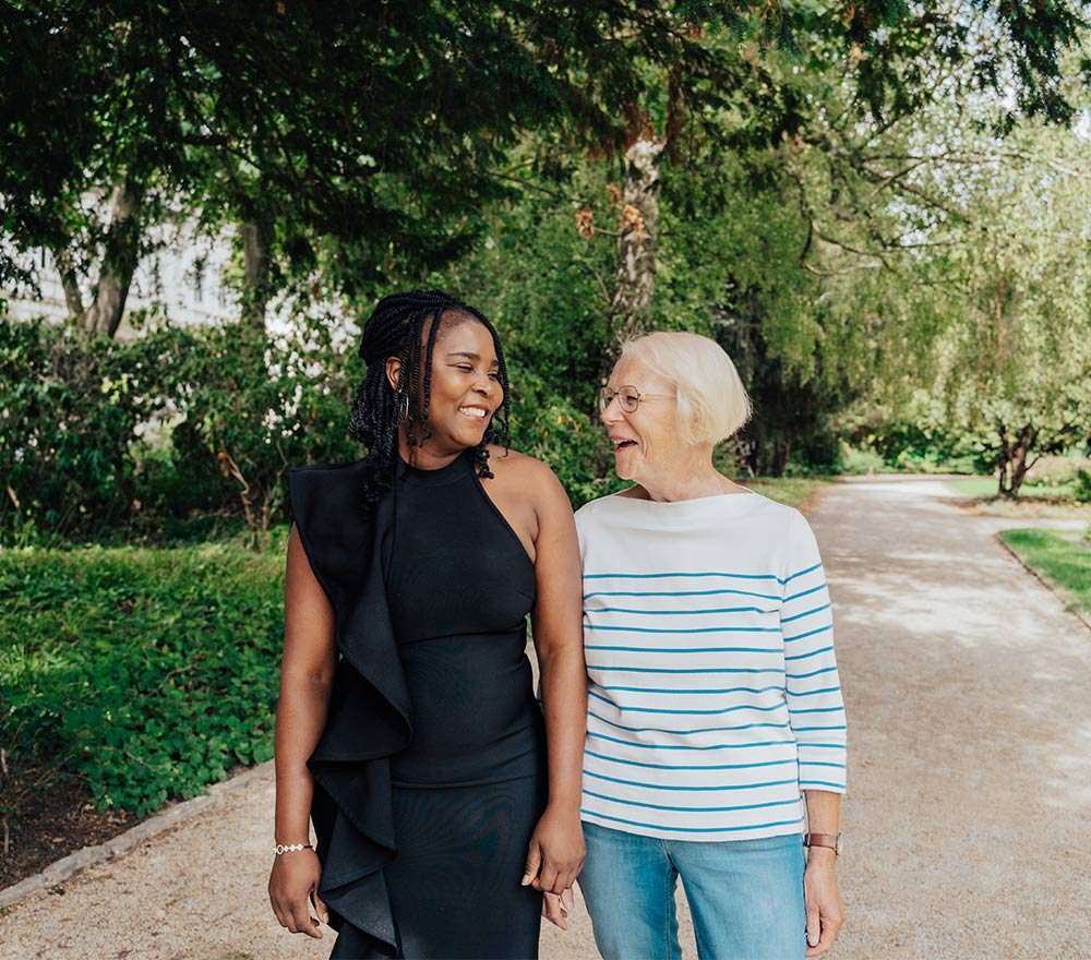 Zwei Frauen – die eine trägt ein schwarzes Kleid, die andere ein gestreiftes Shirt und Jeans – lächeln und gehen gemeinsam auf einem Gartenweg, der von Bäumen und Grün umgeben ist.