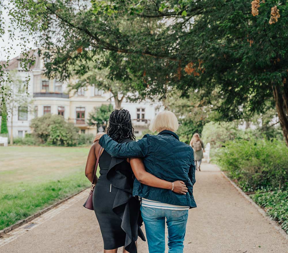 Zwei Personen gehen Arm in Arm einen Parkweg entlang, von hinten gesehen. Die eine trägt ein Kleid, die andere Jeans. Im Hintergrund sind Bäume und Gebäude zu sehen.