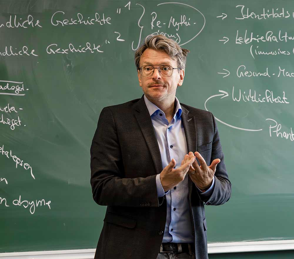 Ein Mann im Anzug steht vor einer mit handschriftlichen Notizen bedeckten Tafel und gestikuliert mit den Händen, während er offenbar einer Klasse etwas erklärt – vielleicht spricht er gerade über das Thema „Generationen in der Kinderliteratur“.