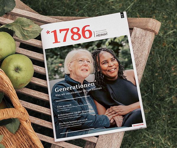 Auf einer Holzbank neben grünen Äpfeln liegt eine Zeitschrift mit dem Titel „1786“. Auf dem Titelblatt sind zwei Frauen zu sehen, eine ältere und eine jüngere, die gemeinsam im Freien lächeln; dazu steht ein deutscher Text über Generationen und das gegenseitige Lernen.