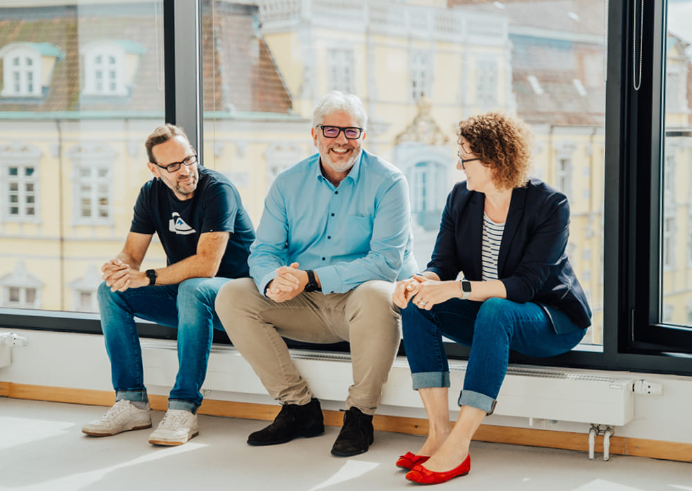 Drei Personen sitzen an einem großen Fenster, lächeln und unterhalten sich. Die Person in der Mitte, die vielleicht über die neuesten Trends im Eventmanagement spricht oder Erkenntnisse aus Florian Zänglers Podcast weitergibt, trägt eine Brille und ein blaues Hemd; die beiden anderen sind leger gekleidet. Draußen sind hell beleuchtete Gebäude zu sehen.