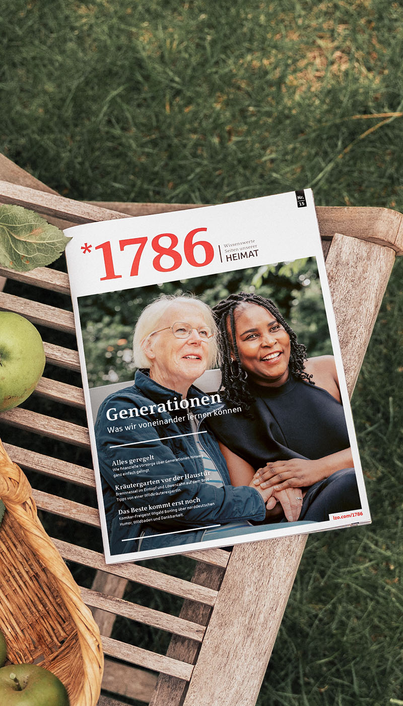 Auf einem Holztisch neben grünen Äpfeln liegt eine Zeitschrift mit dem Titel 1786; auf dem Titelblatt sind zwei lächelnde Frauen zu sehen, eine ältere und eine jüngere, die gemeinsam im Freien sitzen.