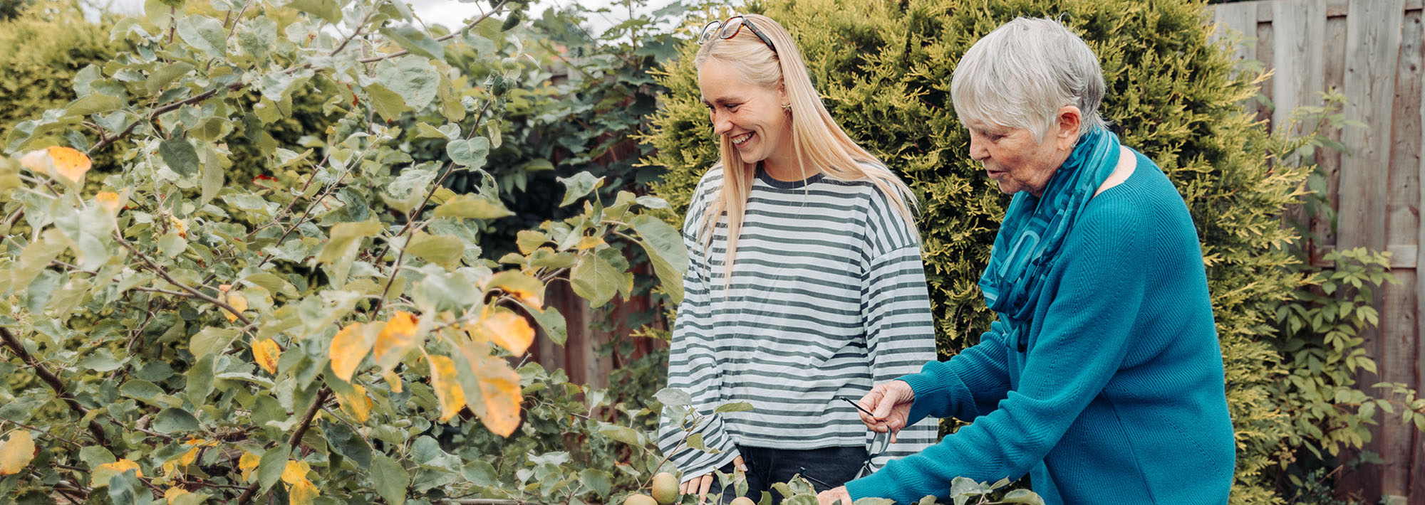 Zwei Frauen, eine ältere und eine jüngere, stehen gemeinsam in einem Garten, der von Grünflächen und Bäumen umgeben ist. Die ältere Frau schneidet Äste zurück, während die jüngere Frau lächelt; beide genießen die Tätigkeit im Freien.