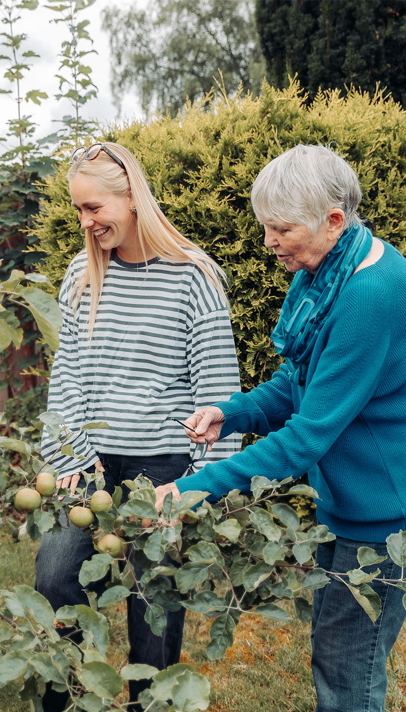Zwei Frauen stehen in einem Garten, lächeln und pflücken Äpfel von einem Baum. Die eine ist jünger, hat langes blondes Haar und trägt ein gestreiftes Shirt, die andere ist älter, hat kurzes graues Haar und trägt einen blauen Pullover. Im Hintergrund ist üppiges Grün zu sehen.