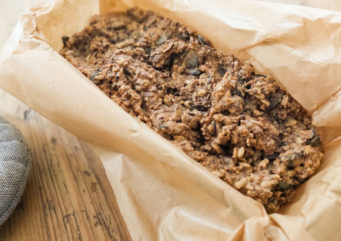 Eine Nahaufnahme eines mit Körnern und Samen gefüllten Brotes, das in einer mit Pergamentpapier ausgelegten Backform auf einer Holzplatte liegt - perfekt für alle, die auf der Suche nach dem besten Rezept für Brote sind.