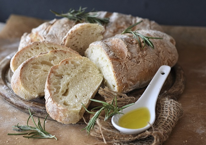 Ein rustikaler Laib geschnittenen Brotes liegt auf einem Holzbrett, garniert mit Rosmarinzweigen. Ein weißer, mit Olivenöl gefüllter Keramiklöffel ruht daneben auf Sackleinen - die perfekte Inspiration für Ihr nächstes Rezept Brote.